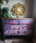A wooden chest of drawers features an intricate floral design with pink and purple roses on the front. Above the chest hangs a decorative, ornate gold clock. A potted plant stands to the left, and the furnishings sit against a light gray wall.