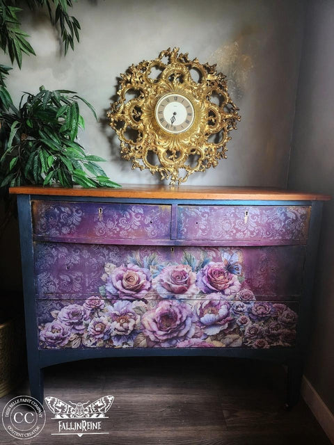 A wooden chest of drawers features an intricate floral design with pink and purple roses on the front. Above the chest hangs a decorative, ornate gold clock. A potted plant stands to the left, and the furnishings sit against a light gray wall.