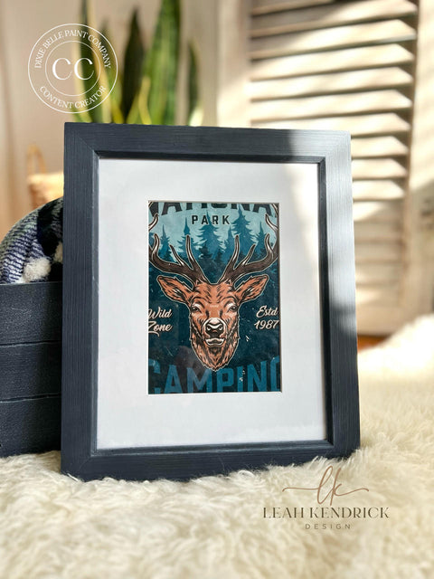A framed artwork featuring National Parks A3 Rice Decoupage Paper with a stylized stag and the words "National Park, Wild Zone, Estd. 1987, Camping" is displayed against a cozy background with a navy blue and white patterned textile and a fluffy white surface, suggesting a homey, rustic decor vibe.
