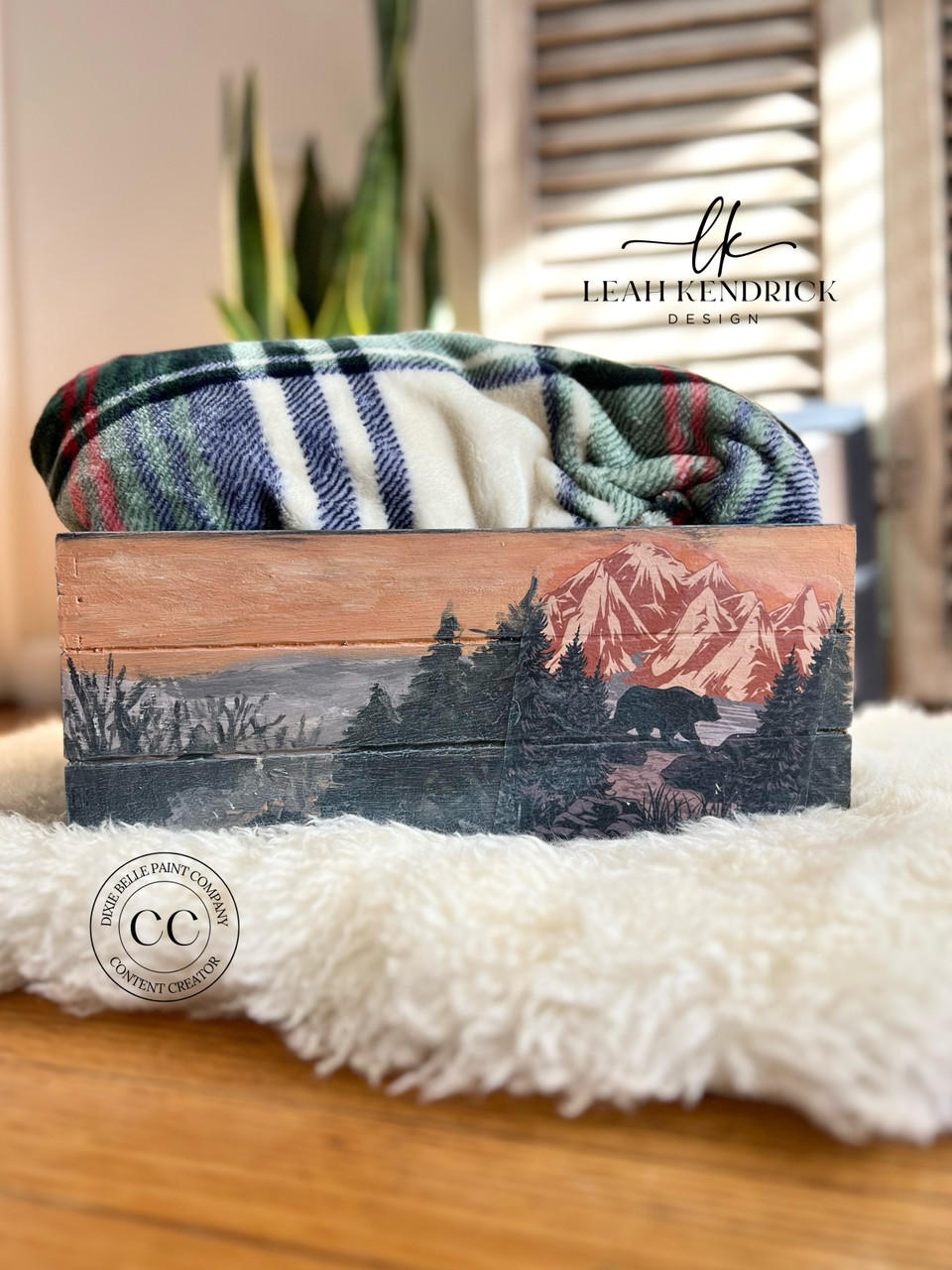 A cozy and artistic scene featuring a beautifully hand-painted wooden box with a mountainous landscape, positioned on a soft, fluffy white surface with a plaid blanket gently draped over it, evoking a sense of warmth and homeliness using the National Parks A3 Rice Decoupage Paper.