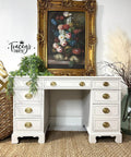 A vintage white desk with brass handles sits beneath a framed floral painting. Greenery in a pot, pampas grass, and a woven basket decorate the scene. "Tracey's Fancy" watermark is shown on the white wall.