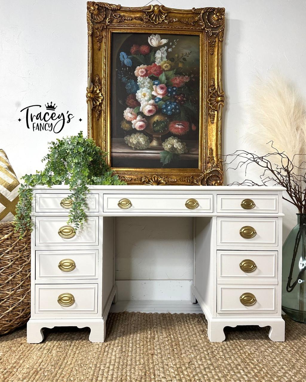 A vintage white desk with brass handles sits beneath a framed floral painting. Greenery in a pot, pampas grass, and a woven basket decorate the scene. "Tracey's Fancy" watermark is shown on the white wall.