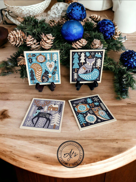 Holiday-themed coasters featuring winter scenes with animals are displayed on a wooden table. Two coasters are standing on small black easels, and two are laid flat. Pine and blue ornament decorations are in the background. A circular logo is present at the bottom.
