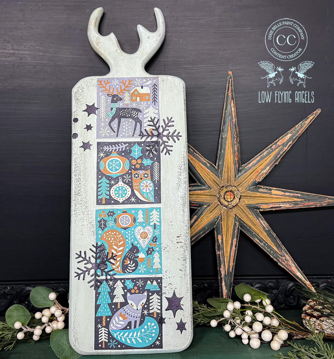A rectangular board with a handle and antler-shaped top displays a vibrant winter-themed design featuring a stag, owl, squirrel, and fox among trees and snowflakes in shades of blue, green, and orange. Nearby is a large rustic star and clusters of white berries.