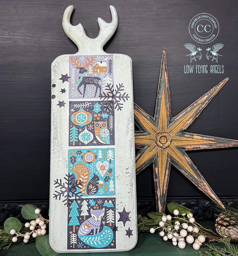 A rectangular board with a handle and antler-shaped top displays a vibrant winter-themed design featuring a stag, owl, squirrel, and fox among trees and snowflakes in shades of blue, green, and orange. Nearby is a large rustic star and clusters of white berries.