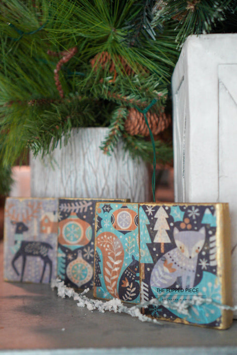 Four decorative Christmas blocks featuring whimsical designs using the Nordic Noel Decoupage Paper, including trees and animals, placed under potted pine trees.