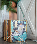 Gold blocks wrapped adorned with the Nordic Noel Decoupage Paper with snowflakes and trees, tied with a white string. Background features subtle, rustic decor including a wooden surface and green pine branches.