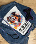 A denim jacket with a colorful print of a happy dog and the text "RESCUED IS MY FAVORITE BREED" on the back, displayed on a wooden floor. Next to the jacket are a black bone-shaped dog toy with purple edges and a rainbow-colored leash.
