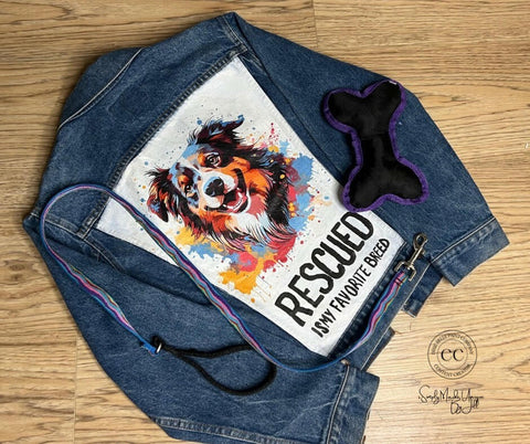 A denim jacket with a colorful print of a happy dog and the text "RESCUED IS MY FAVORITE BREED" on the back, displayed on a wooden floor. Next to the jacket are a black bone-shaped dog toy with purple edges and a rainbow-colored leash.