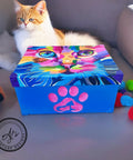 A fluffy, orange and white cat lounges on a gray surface in front of a brightly colored, painted box. The box has a vibrant cat face on the lid and a pink paw print with a dog and cat silhouette inside on the front. Several colorful pom-pom toys are scattered next to it.