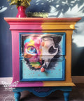 A colorful painted dresser features a vibrant cat design split between two drawers. The left side of the cat is bright and psychedelic, while the right side is monochrome and skeletal. The dresser is set against a blue wall, with a vase of sunflowers on top.