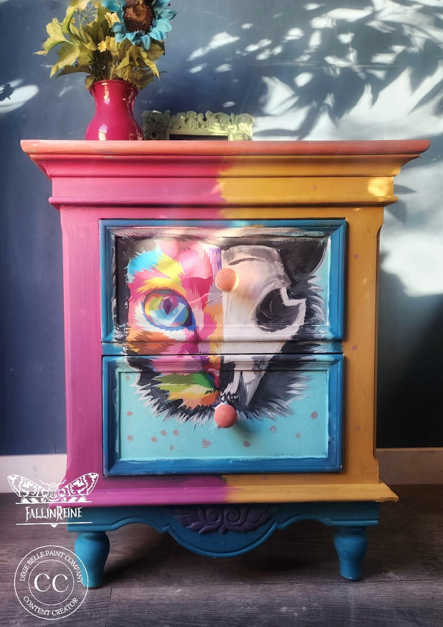 A colorful painted dresser features a vibrant cat design split between two drawers. The left side of the cat is bright and psychedelic, while the right side is monochrome and skeletal. The dresser is set against a blue wall, with a vase of sunflowers on top.