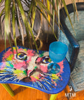 A colorful table featuring a vibrant, abstract painting of a cat's face stands next to a potted plant with long, narrow leaves. A bright blue goblet sits on the table. A teal and navy chair is placed nearby.