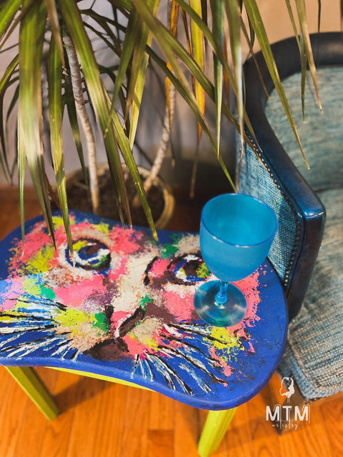 A colorful table featuring a vibrant, abstract painting of a cat's face stands next to a potted plant with long, narrow leaves. A bright blue goblet sits on the table. A teal and navy chair is placed nearby.