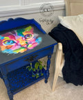A small, ornate blue table with a colorful, abstract painting of a cat's face on its top. The bottom shelf has a blue leopard print design and holds a small plant. The table is next to a beige armchair with a white pillow and a fluffy black blanket draped over it.