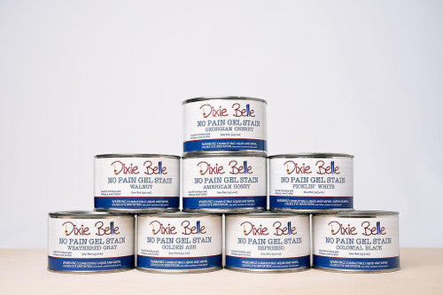 Eight cans of Dixie Belle No Pain Gel Stain are stacked in a pyramid on a wooden surface. Visible stain colors include Georgian Cherry, Walnut, American Honey, Picklin' White, Weathered Gray, Golden Ash, Espresso, and Colonial Black.