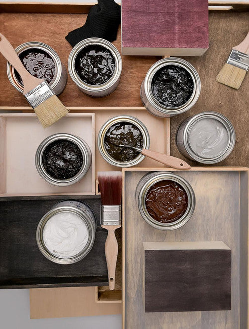 Open cans of wood stain in various colors, with paint brushes resting on the lids and trays, are arranged on a wooden surface alongside stained wood samples in different shades.