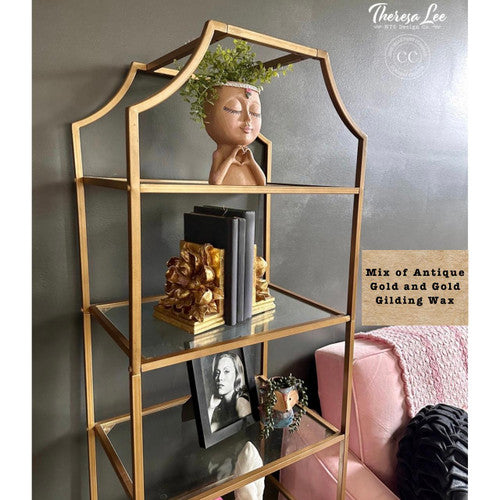 A gold and glass shelving unit holds a plant in a face-shaped pot, gold decorative pieces, books, and a framed black-and-white photograph. The shelves are next to a pink sofa against a gray wall. Text reads: “Mix of Antique Gold and Gold Gilding Wax.”.