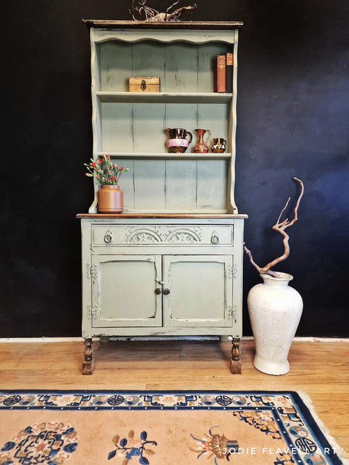A vintage green cabinet with ornate details stands against a dark wall. It displays a decorative box, books, and shiny cups. Below, a large white vase with twisted branches sits to the right. A rug with floral patterns lies on the wooden floor.