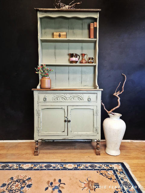 A vintage green cabinet with ornate details stands against a dark wall. It displays a decorative box, books, and shiny cups. Below, a large white vase with twisted branches sits to the right. A rug with floral patterns lies on the wooden floor.