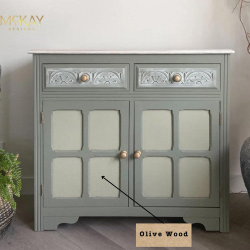 A light green cabinet with intricate patterns on the top drawers and gold knobs. "Olive Wood" is labeled with an arrow pointing to a section. The cabinet stands on a wooden floor with a potted plant nearby.