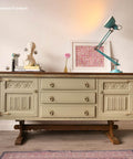 Vintage sideboard with intricate detailing, featuring three central drawers, flanked by two cabinets. On top are a white bust, a pink flower in a vase, a turquoise lamp, and a framed abstract art piece, set against a white wall.