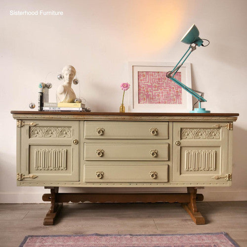 Vintage sideboard with intricate detailing, featuring three central drawers, flanked by two cabinets. On top are a white bust, a pink flower in a vase, a turquoise lamp, and a framed abstract art piece, set against a white wall.