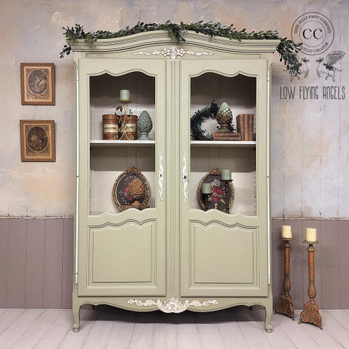 A vintage green cabinet with glass-paneled doors displays decorative plates, books, and ornaments. It’s topped with greenery. The wall features framed art and a watermark for “Low Flying Angels” with a winged design and “Dixie Belle Paint Company Content Creator” . Candlesticks sit nearby.