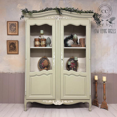 A vintage green cabinet with glass-paneled doors displays decorative plates, books, and ornaments. It’s topped with greenery. The wall features framed art and a watermark for “Low Flying Angels” with a winged design and “Dixie Belle Paint Company Content Creator” . Candlesticks sit nearby.