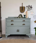 A light green vintage sideboard with brass handles is decorated with a candle, greenery, and a woven wall hanging. Plants and a ladder with a blue blanket are nearby, creating a cozy, stylish interior scene.