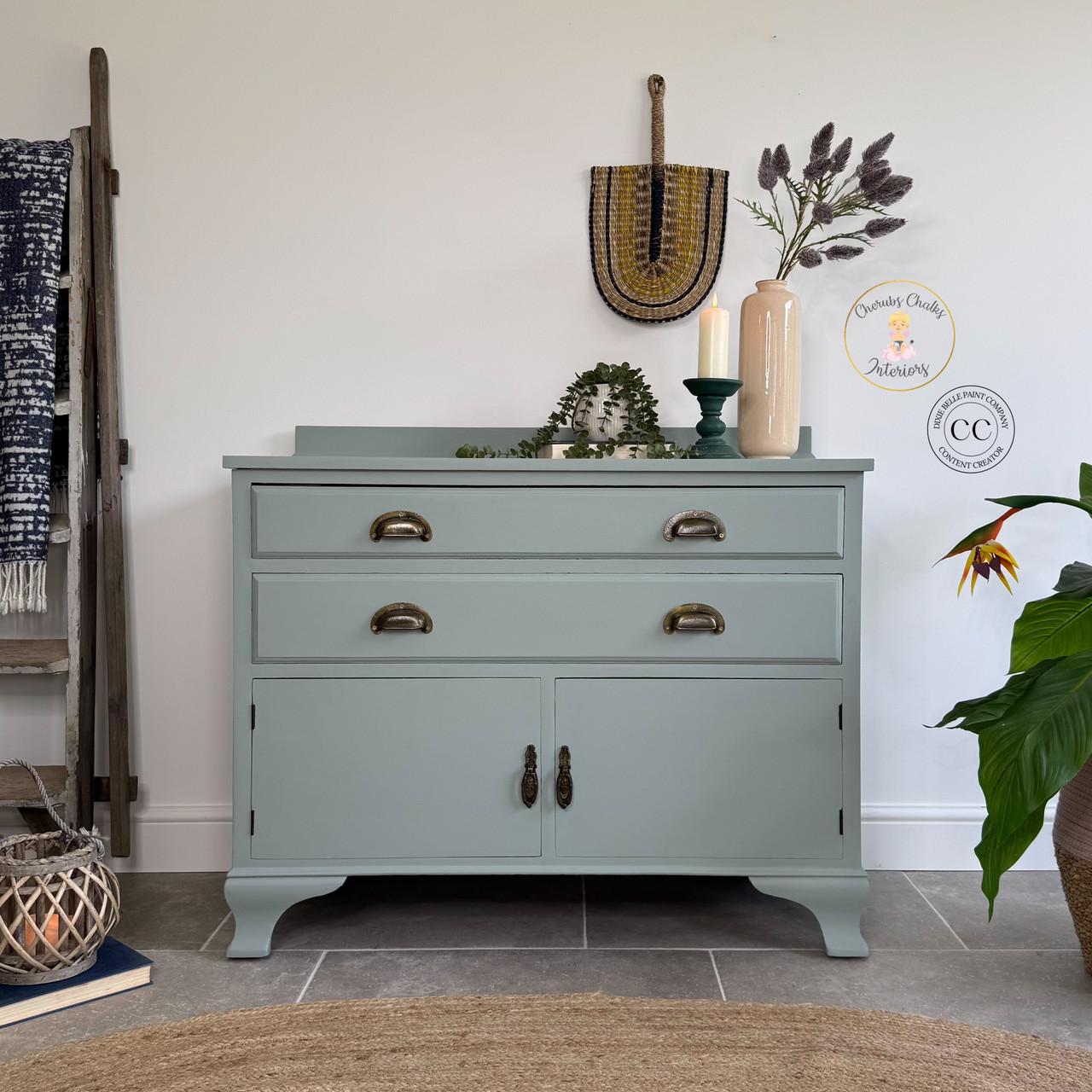 A light green vintage sideboard with brass handles is decorated with a candle, greenery, and a woven wall hanging. Plants and a ladder with a blue blanket are nearby, creating a cozy, stylish interior scene.