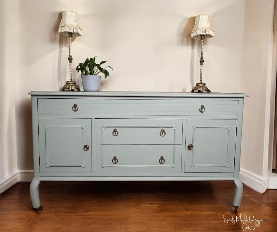 A light blue vintage sideboard with brass handles stands against a cream wall, flanked by two ornate table lamps with white shades. A small potted plant sits on top, and the floor is wooden.