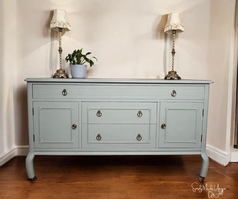 A light blue vintage sideboard with brass handles stands against a cream wall, flanked by two ornate table lamps with white shades. A small potted plant sits on top, and the floor is wooden.