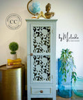 A light blue cabinet with intricate floral cut-out doors stands against a wall, topped with a globe and decorative items. Next to it is a brass table with a plant, and a large glass vase with greenery on the floor.