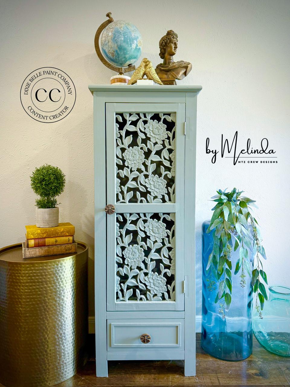 A light blue cabinet with intricate floral cut-out doors stands against a wall, topped with a globe and decorative items. Next to it is a brass table with a plant, and a large glass vase with greenery on the floor.