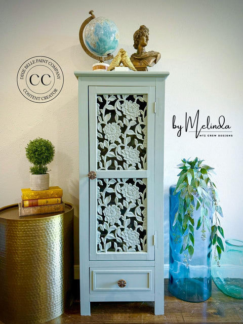 A light blue cabinet with intricate floral cut-out doors stands against a wall, topped with a globe and decorative items. Next to it is a brass table with a plant, and a large glass vase with greenery on the floor.