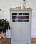 A light blue vintage cabinet with a glass-front upper door and decorative molding on the lower door stands on a wooden floor next to a potted plant. Candles and decor are arranged on top against a white brick wall.