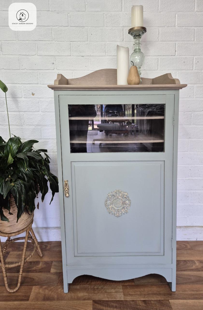 A light blue vintage cabinet with a glass-front upper door and decorative molding on the lower door stands on a wooden floor next to a potted plant. Candles and decor are arranged on top against a white brick wall.