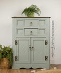 A vintage mint-green cabinet with two drawers and two doors, featuring ornate metal handles and decorative carvings, stands on a wooden floor with green plants on and beside it. A watermark reads â€œGRACIEâ€™S HOUSE Painted Furniture.â€.