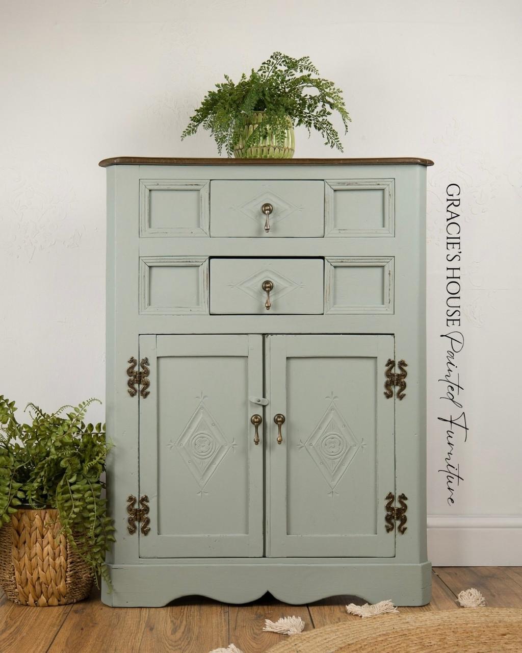 A vintage mint-green cabinet with two drawers and two doors, featuring ornate metal handles and decorative carvings, stands on a wooden floor with green plants on and beside it. A watermark reads â€œGRACIEâ€™S HOUSE Painted Furniture.â€.
