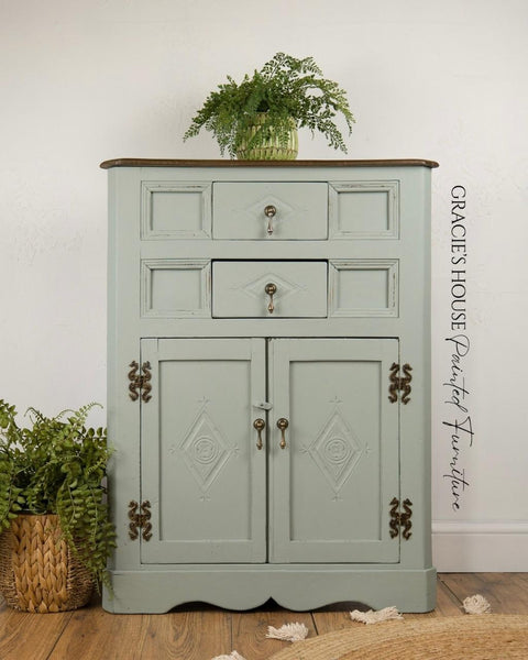 A vintage mint-green cabinet with two drawers and two doors, featuring ornate metal handles and decorative carvings, stands on a wooden floor with green plants on and beside it. A watermark reads â€œGRACIEâ€™S HOUSE Painted Furniture.â€.