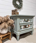 A light blue, vintage-style nightstand with floral designs on the drawers sits against a white shiplap wall. A leafy wreath hangs above. Dried plants in vases and wooden decor items are arranged beside and on top of the nightstand.