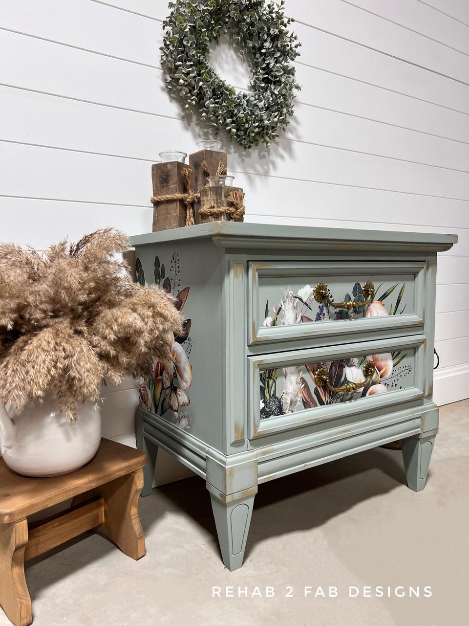 A light blue, vintage-style nightstand with floral designs on the drawers sits against a white shiplap wall. A leafy wreath hangs above. Dried plants in vases and wooden decor items are arranged beside and on top of the nightstand.