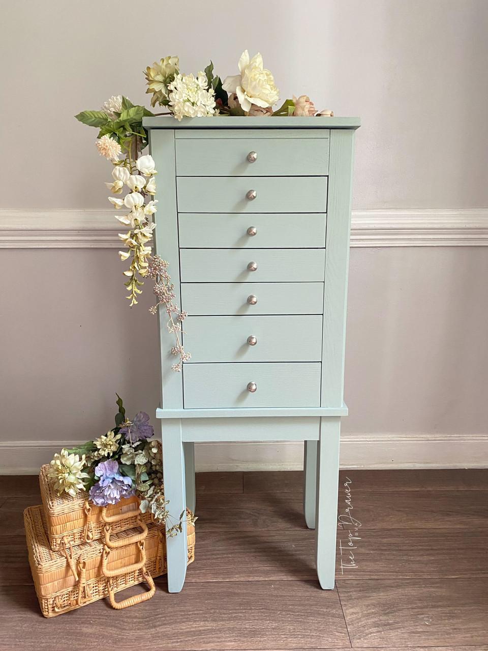A tall, light blue wooden chest with seven drawers stands against a beige wall, decorated with white and pale flowers on top. Wicker baskets with more flowers are arranged on the floor beside it.
