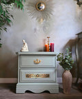 A small sage green dresser with two drawers sits against a gray wall. On top are a white statue, two lit candles, and a gold candlestick. A round sunburst mirror and leafy plant decor are in the background. A large vase with leaves is beside the dresser.