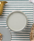 A top-down view of an open can of light greenish blue paint on a striped white and green surface, surrounded by decorative items including a gold object, a round gray sample, and a metallic tray.