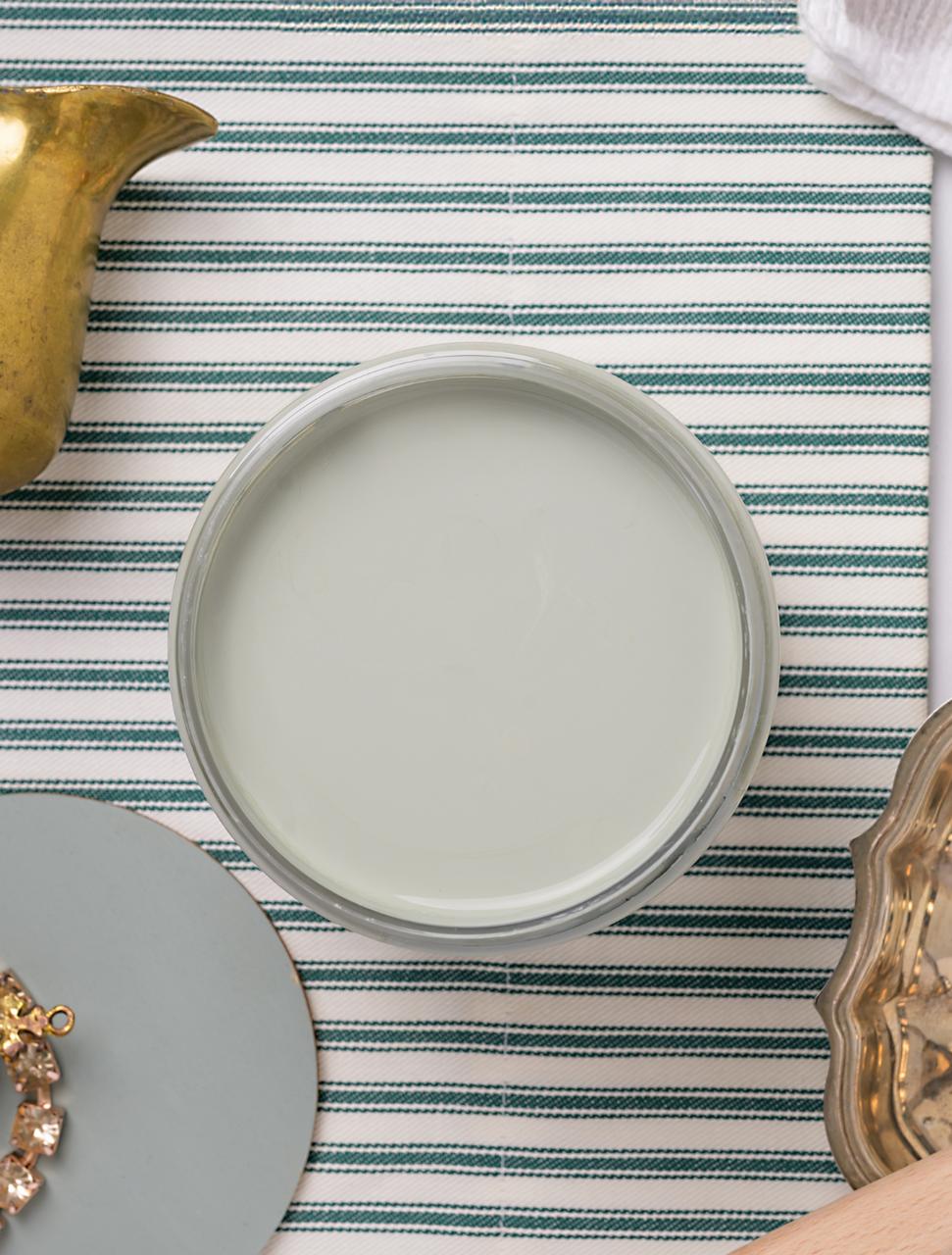 A top-down view of an open can of light greenish blue paint on a striped white and green surface, surrounded by decorative items including a gold object, a round gray sample, and a metallic tray.