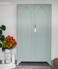 A light green vintage wardrobe stands against a white wall. Next to it is a potted Monstera plant, a white vase with orange flowers, and a sculpted face vase. A white tassel hangs from the wardrobe handle.