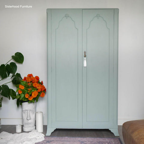 A light green vintage wardrobe stands against a white wall. Next to it is a potted Monstera plant, a white vase with orange flowers, and a sculpted face vase. A white tassel hangs from the wardrobe handle.