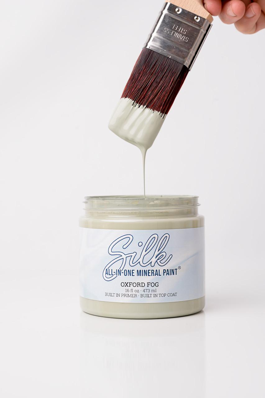 A hand holds a paintbrush dripping light grey paint over an open jar labeled "Silk All-In-One Mineral Paint Oxford Fog" against a plain white background.